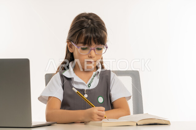 متابعة الحصص و حل الواجبات عبر الانترنت، طالبة مجتهدة تراجع دروسها من الكتب وتستخدم الحاسوب المحمول، بورتريه مقرب لتلميذة خليجية عربية إماراتية ترتدي اليونيفورم المدرسي و تدرس في المنزل، المذاكرة و الدراسة عن بعد، مفهوم التعليم الإلكتروني، خلفية بيضاء