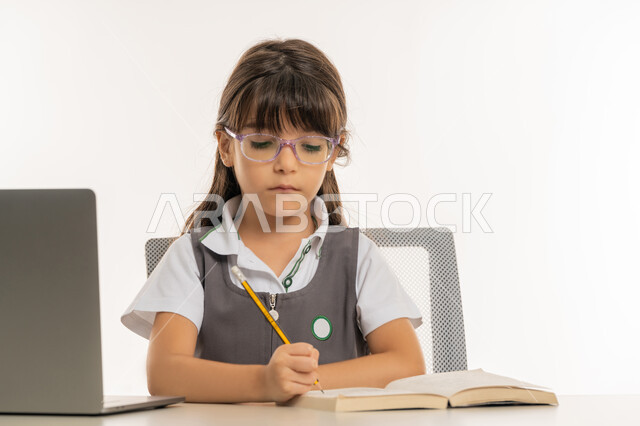 طالبة مجتهدة تراجع دروسها من الكتب وتستخدم الحاسوب المحمول، متابعة الحصص و حل الواجبات عبر الانترنت، بورتريه مقرب لتلميذة خليجية عربية إماراتية ترتدي اليونيفورم المدرسي و تدرس في المنزل، المذاكرة و الدراسة عن بعد، مفهوم التعليم الإلكتروني، خلفية بيضاء