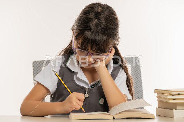 ايماءات الاندماج والتركيز، القيام بالمهام التعليمية الفعالة, بورتريه مقرب لطالبة عربية خليجية إماراتية ترتدي المريول المدرسي وتجلس على طاولة الدراسة, تلميذة مجتهدة تقوم بتدوين المعلومات لتمكين وفهم الدروس, اكتساب المعرفة من كتابة الواجبات، خلفية بيضاء