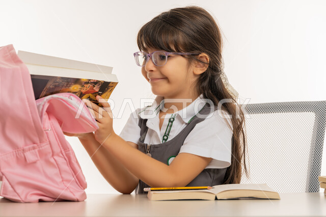 تجهيز الطاولة الدراسية لمراجعة الدروس، الاجتهاد والتفوق ومتابعة الدراسة، بورتريه لتلميذة عربية خليجية اماراتية ترتدي اليونيفورم المدرسي تقوم بسحب الكتب من الحقيبة، طالبة مجتهدة متحمسة للقراءة والحفظ وإنهاء الواجبات المنزلية المطلوبة، خلفية بيضاء