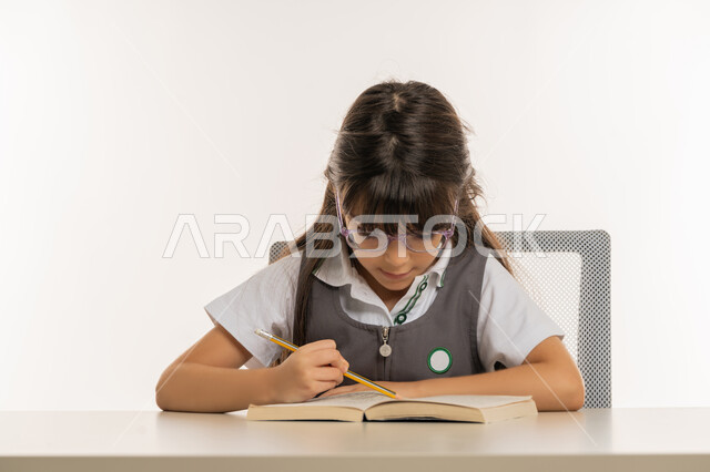 القيام بالمهام التعليمية الفعالة, بورتريه مقرب لطالبة عربية خليجية إماراتية ترتدي المريول المدرسي وتجلس على طاولة الدراسة, تلميذة مجتهدة تقوم بتدوين المعلومات لتمكين وفهم الدروس, ايماءات الاندماج والتركيز، اكتساب المعرفة من كتابة الواجبات، خلفية بيضاء