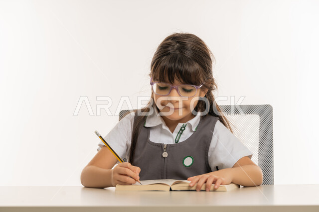 اكتساب المعرفة من كتابة الواجبات، القيام بالمهام التعليمية الفعالة, بورتريه مقرب لطالبة عربية خليجية إماراتية ترتدي المريول المدرسي وتجلس على طاولة الدراسة, تلميذة مجتهدة تقوم بتدوين المعلومات لتمكين وفهم الدروس, ايماءات الاندماج والتركيز، خلفية بيضاء