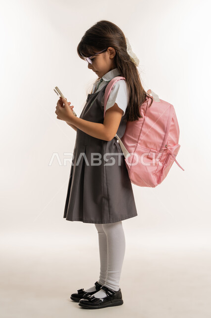 استخدام التكنولوجيا في التعليم، متابعة الدروس عن بعد, بورتريه لطالبة عربية خليجية إماراتية ترتدي اليونيفورم المدرسي وحقيبة الظهر تستخدم الهاتف المحمول في الدراسة وحل الواجبات, تلميذة مجتهدة تقوم بإجراء بحث عبر الانترنت, خلفية بيضاء