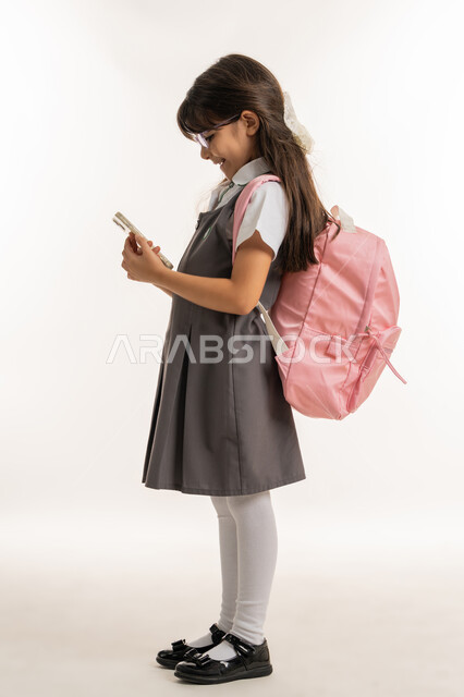 مشاركة مشاعر السرور لبدء العام الدراسي, بورتريه لطالبة عربية خليجية إماراتية ترتدي اليونيفورم المدرسي وحقيبة الظهر  تستخدم الهاتف المحمول في الدراسة وحل الواجبات, تلميذة مجتهدة تقوم بإجراء بحث عبر الانترنت, استخدام التكنولوجيا في التعليم، خلفية بيضاء