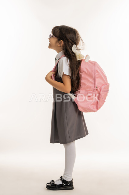 إيماءات تدل على الحماس والسعادة، الرغبة في طلب العلم، موسم العوده للمدارس, التهيئة والتجهيز لبداية العام الدراسي, بورتريه من الجانب لطالبة عربية خليجية اماراتية ترتدي اليونيفورم الخاص وتحمل الحقيبة المدرسية, تلميذه مجتهده ومتميزه, خلفية بيضاء.