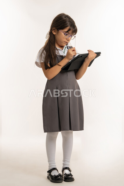 مفهوم التعليم الالكتروني عبر جهاز التابليت, بورتريه لتلميذة عربية خليجية إماراتية ترتدي اليونيفورم المدرسي وحقيبة الظهر تمسك بيدها الآيباد، طالبة سعودية تقوم بالبحث عن معلومات لحل الواجبات المدرسية، متابعة الدروس عن بعد عن طريق الانترنت, خلفية بيضاء