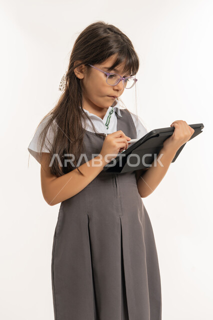 متابعة الدروس عن بعد عن طريق الانترنت, مفهوم التعليم الالكتروني عبر جهاز التابليت, بورتريه لتلميذة عربية خليجية إماراتية ترتدي اليونيفورم المدرسي وحقيبة الظهر تمسك بيدها الآيباد، طالبة سعودية تقوم بالبحث عن معلومات لحل الواجبات المدرسية، خلفية بيضاء