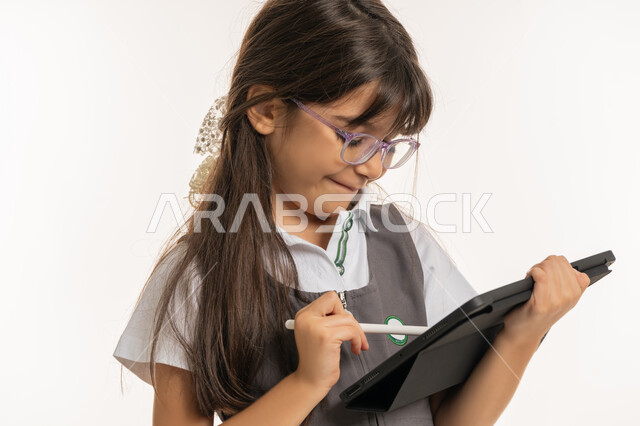 طالبة سعودية تقوم بالبحث عن معلومات لحل الواجبات المدرسية، متابعة الدروس عن بعد عن طريق الانترنت, مفهوم التعليم الالكتروني عبر جهاز التابليت, بورتريه مقرب لتلميذة عربية خليجية إماراتية ترتدي اليونيفورم المدرسي وحقيبة الظهر تمسك بيدها الآيباد، خلفية بيضاء