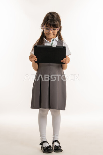 E-learning concept via tablet, portrait of an Arab Gulf Emirati student wearing school uniform and backpack holding iPad in hand, Saudi student searching for information to solve homework, following lessons remotely via the internet, white background