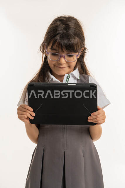 Following lessons remotely via the Internet, the concept of e-learning via a tablet, a portrait of an Arab Gulf Emirati student wearing a school uniform and a backpack holding an iPad in her hand, a Saudi student searching for information to solve school assignments, white background