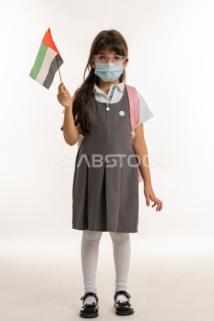 العوده بحذر للدراسه الحضوريه، الإحتفال وإحياء ذكرى اليوم الوطني الاماراتي في المدارس، بورتريه لطالبه عربية خليجية اماراتية ترتدي الزي المدرسي والحقيبة المدرسية، تلميذه مجتهدة ترتدي الكمامة للوقاية من فايروس كورونا تحمل بيدها علم الامارات، خلفية بيضاء.