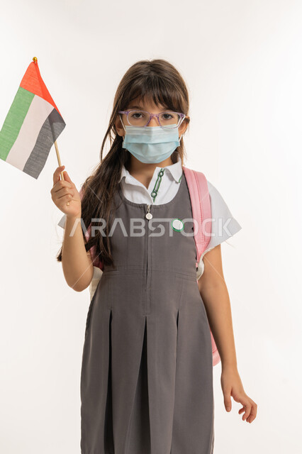 تلميذه مجتهدة ترتدي الكمامة للوقاية من فايروس كورونا تحمل بيدها علم الامارات، العوده بحذر للدراسه الحضوريه، الإحتفال وإحياء ذكرى اليوم الوطني الاماراتي في المدارس، بورتريه لطالبه عربية خليجية اماراتية ترتدي الزي المدرسي والحقيبة المدرسية، خلفية بيضاء.