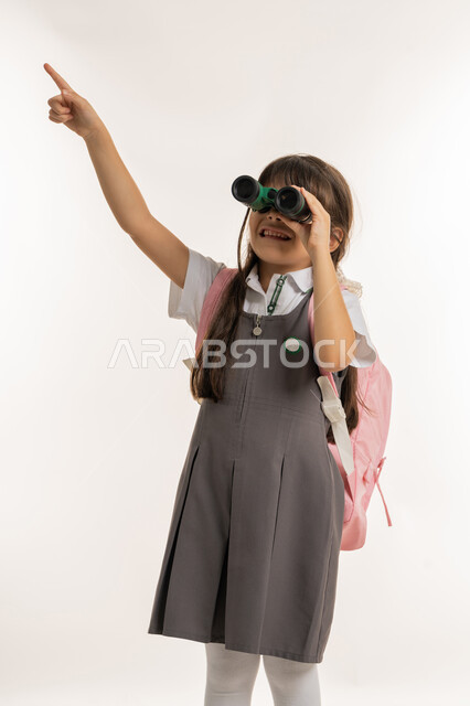 مفهوم التدقيق والتركيز، التخطيط المستقبلي للأهداف والطموحات، حب الاستكشاف والمغامرة، رؤية الأشياء البعيدة باستخدام التلسكوب، بورتريه لطالبة إماراتية خليجية عربية ترتدي اليونيفورم والحقيبة المدرسية وتنظر من خلال المنظار وتشير بيدها الى شيء ما، الخلفية البيضاء
