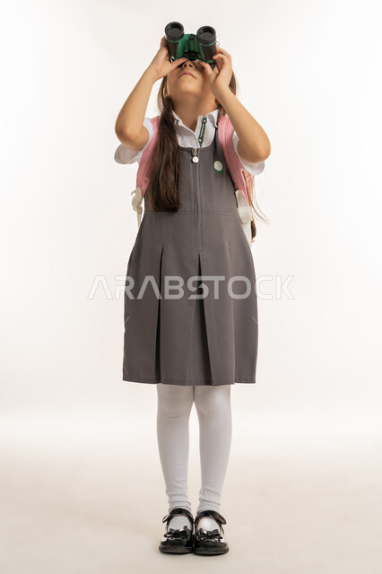 رؤية الأشياء البعيدة باستخدام التلسكوب، بورتريه لطالبة إماراتية خليجية عربية ترتدي اليونيفورم والحقيبة المدرسية وتنظر من خلال المنظار ثنائي العينين، مفهوم التدقيق والتركيز، التخطيط المستقبلي للأهداف والطموحات، حب الاستكشاف والمغامرة، الخلفية البيضاء