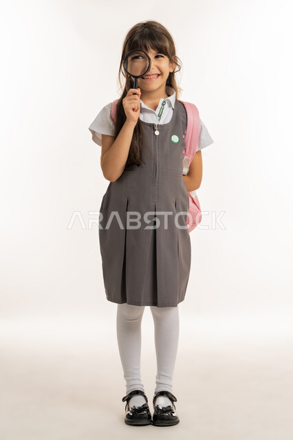 المساعدة على الاستكشاف والبحث، مفهوم التدقيق والتركيز وتوضيح الصورة، الاستعداد للفصل الدراسي الجديد، أهمية التعليم في الإمارات العربية المتحدة، تلميذة عربية خليجية إماراتية ترتدي الزي المدرسي والحقيبة المدرسية تستخدم عدسة مكبرة، خلفية بيضاء