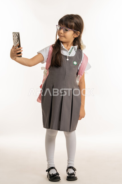 الاستعداد للذهاب للمدرسة وبداية العام الجديد, بورتريه لتلميذة عربية خليجية إماراتية ترتدي المريول المدرسي وحقيبة الظهر وتمسك الهاتف المحمول, طالبة مبتسمة تقوم بالتقاط صور سيلفي تذكارية عبر الجوال, ايماءات الحماس والسرور لعودة المدارس, خلفية بيضاء