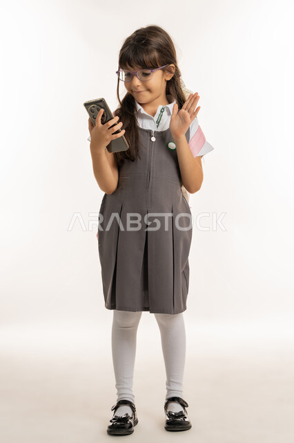 عام دراسي جديد ومخطط تعليمي حديث, بورتريه لطالبة عربية خليجية إمارتية مبتسمة ترتدي المريول المدرسي وحقيبة الظهر وتمسك الهاتف المحمول, تلميذة تقوم بالتواصل الاجتماعي وإجراء مكالمة فيديو عبر الجوال, استخدام الأجهزة المتطورة لدى الأطفال, خلفية بيضاء