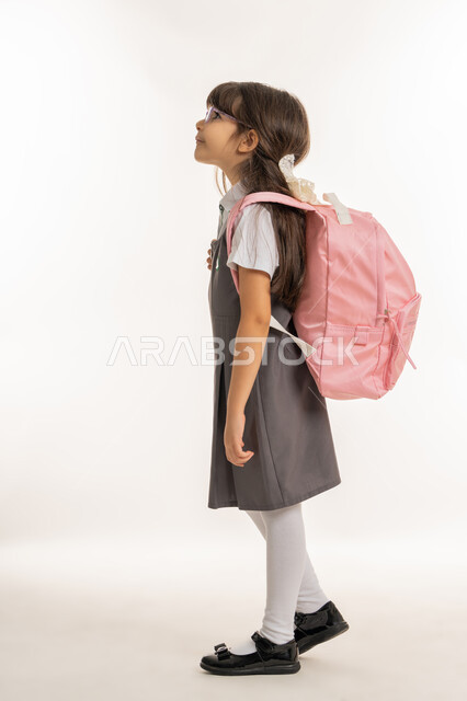 موسم العوده للمدارس, التهيئة والتجهيز لبداية العام الدراسي, بورتريه من الجانب لطالبة عربية خليجية اماراتية ترتدي الزي المدرسي وتحمل الحقيبة المدرسية, تلميذه مجتهده ومتميزه, ايماءات المشي إلى المدرسه, صورة الجسم بالطول الكامل, خلفية بيضاء.