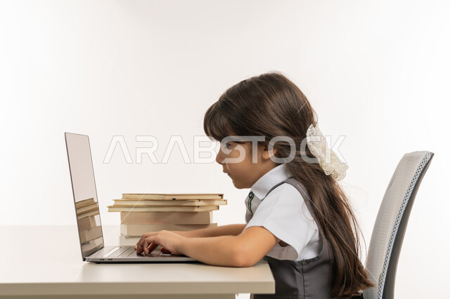 طالبه مجتهده متفوقه تدرس عن بعد عبر الانترنت، الاستعداد للإمتحانات والاختبارات المدرسيه، مفهوم المثابره على التقدم والنجاح، بورتريه مقرب من الجانب لتلميذة خليجية عربية إماراتية ترتدي اليونيفورم المدرسي وتتابع حصص الاونلاين عبر الحاسوب، خلفية بيضاء