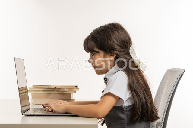 مفهوم المثابره على التقدم والنجاح، بورتريه مقرب من الجانب لتلميذة خليجية عربية إماراتية ترتدي اليونيفورم المدرسي وتتابع حصص الاونلاين عبر الحاسوب، طالبه مجتهده متفوقه تدرس عن بعد عبر الانترنت، الاستعداد للإمتحانات والاختبارات المدرسيه، خلفية بيضاء