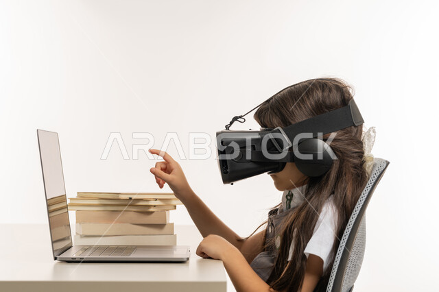 متابعة حصص الاونلاين عن طريق الكمبيوتر، مفهوم التعليم الإلكتروني، طفلة مجتهدة ترتدي نظارات الواقع الإفتراضي وتتفاعل مع البث المباشر، حل الواجبات عبر الانترنت، بورتري مقرب من الجانب لتلميذة خليجية عربية إماراتية ترتدي اليونيفورم و تشير الى شيئ ما، خلفية بيضاء