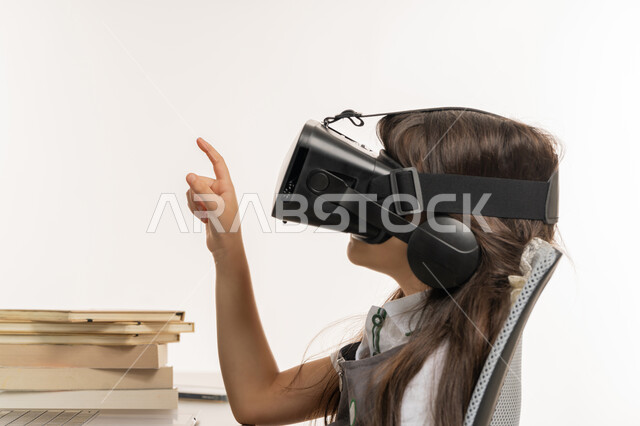 حل الواجبات عبر الانترنت، بورتري مقرب من الجانب لتلميذة خليجية عربية إماراتية ترتدي اليونيفورم و تشير الى شيئ ما، متابعة حصص الاونلاين عن طريق الكمبيوتر، مفهوم التعليم الإلكتروني، طفلة مجتهدة ترتدي نظارات الواقع الإفتراضي وتتفاعل مع البث المباشر، خلفية بيضاء