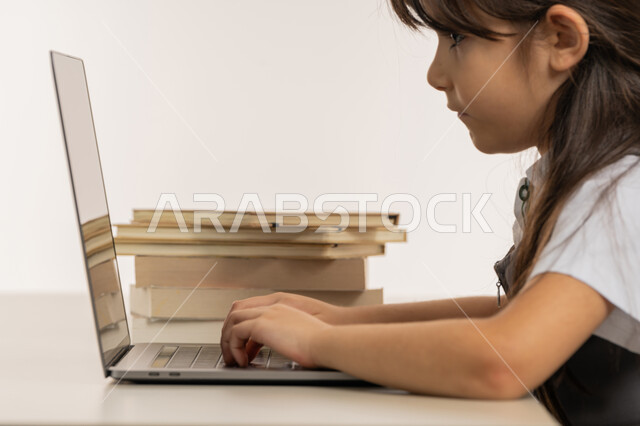 Studying and distance learning, the concept of e-learning, a diligent student reviews her lessons from books and uses a laptop, following up on classes and solving homework via the Internet, a close-up portrait of an Arab Gulf Emirati student wearing a school uniform and studying at home, white background