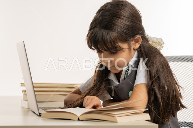 متابعة الحصص و حل الواجبات عبر الانترنت، بورتريه مقرب لتلميذة خليجية عربية إماراتية ترتدي اليونيفورم المدرسي و تدرس في المنزل، المذاكرة و الدراسة عن بعد، مفهوم التعليم الإلكتروني، طالبة مجتهدة تراجع دروسها من الكتب وتستخدم الحاسوب المحمول، خلفية بيضاء