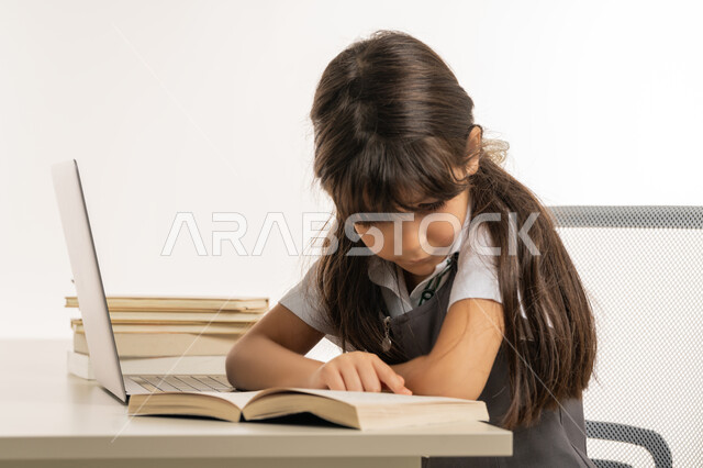 E-learning concept, diligent student reviews her lessons from books and uses laptop, following classes and doing homework online, close-up portrait of an Arab Gulf Emirati student wearing school uniform and studying at home, studying and distance learning, white background