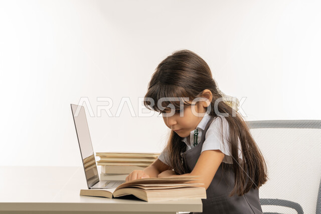 المذاكرة و الدراسة عن بعد، مفهوم التعليم الإلكتروني، طالبة مجتهدة تراجع دروسها من الكتب وتستخدم الحاسوب المحمول، متابعة الحصص و حل الواجبات عبر الانترنت، بورتريه مقرب لتلميذة خليجية عربية إماراتية ترتدي اليونيفورم المدرسي و تدرس في المنزل، خلفية بيضاء