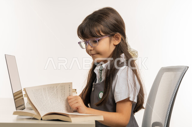 المذاكرة و الدراسة عن بعد، مفهوم التعليم الإلكتروني، طالبة مجتهدة تراجع دروسها من الكتب وتستخدم الحاسوب المحمول، متابعة الحصص و حل الواجبات عبر الانترنت، بورتريه مقرب لتلميذة خليجية عربية إماراتية ترتدي اليونيفورم المدرسي و تدرس في المنزل، خلفية بيضاء