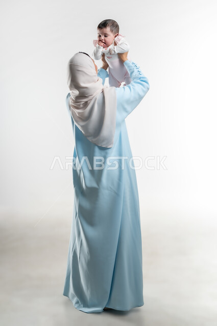 النظر إلى المولوده الرضيعه بإيماءات الحنان والحب, والدة سعودية تقوم بالاستمتاع بقضاء وقت ممتع مع الأطفال, بورتريه من الخلف لأم عربية خليجيه عمانية ترتدي عباءة ملونة تداعب طفلتها, دور الأهل في تنميه مهارات الأطفال الحسيه والحركيه, صورة الجسم بالطول الكامل, خلفيه بيضاء