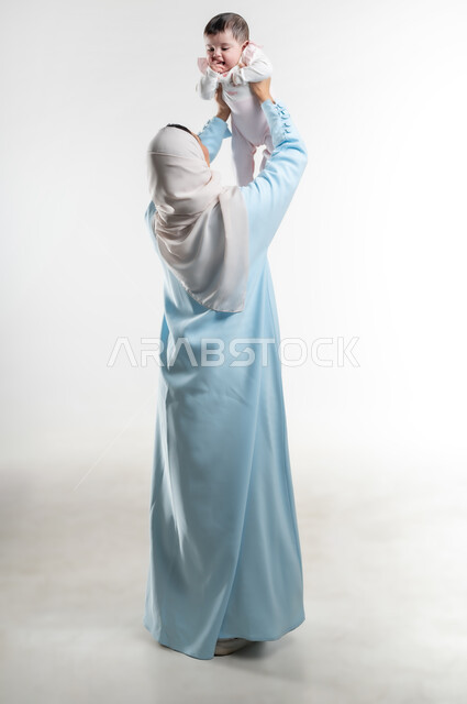 الاستمتاع بقضاء وقت ممتع مع الأطفال, والدة سعودية تقوم بالنظر إلى المولوده الرضيعه بإيماءات الحنان والحب, بورتريه من الخلف لأم عربية خليجيه عمانية ترتدي عباءة ملونة تداعب طفلتها, دور الأهل في تنميه مهارات الأطفال الحسيه والحركيه, صورة الجسم بالطول الكامل, خلفيه بيضاء