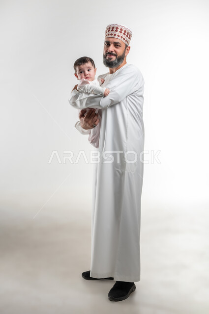 ايماءات الحب والعطف على الصغار, بورتريه لأب عربي خليجي عماني يرتدي الدشداشة ويضع الكمة يحمل ابنته الرضيعة بيديه, قضاء وقت ممتع مع الاطفال,  صورة الجسم بالطول الكامل, خلفية بيضاء