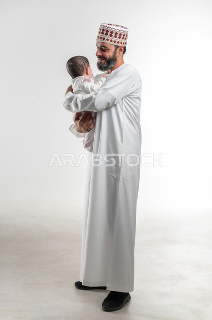 ايماءات الحب والعطف على الصغار, بورتريه لأب عربي خليجي عماني يرتدي الدشداشة ويضع الكمة يحمل ابنته الرضيعة بيديه, قضاء وقت ممتع مع الاطفال,  صورة الجسم بالطول الكامل, خلفية بيضاء