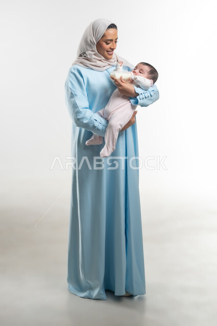 الرعايه والاهتمام بصحة ونمو الأبناء, شرب الحليب من عبوة الرضاعة، مفهوم الأمومة والرضاعة الصناعية، بورتريه مقرب لعائلة عربية خليجية عمانية مع طفلتهم، والدة سعودية تقوم بقضاء أوقات ممتعه مع الأطفال, صورة الجسم بالطول الكامل, خلفية بيضاء