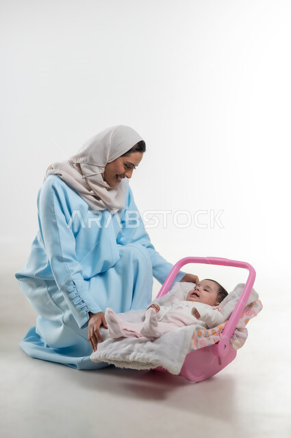 مفهوم الأمومة وتحمل المسؤولية والاهتمام بالأبناء, جلوس الرضيعة على كرسيها الخاص, بورتريه لسيدة عربية خليجيه عمانية تجلس بجانب طفلتها وترتدي الحجاب والعباءة الزرقاء,  والدة سعودية تقوم بقضاء اوقات ممتعة مع الاطفال, خلفية بيضاء