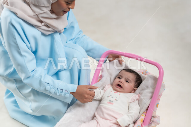 مفهوم الأمومة وتحمل المسؤولية والاهتمام بالأبناء, جلوس الرضيعة على كرسيها الخاص, بورتريه لسيدة عربية خليجيه عمانية تجلس بجانب طفلتها وترتدي الحجاب والعباءة الزرقاء,  والدة سعودية تقوم بقضاء اوقات ممتعة مع الاطفال, خلفية بيضاء