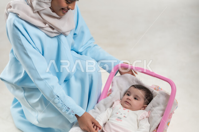 مفهوم الأمومة وتحمل المسؤولية والاهتمام بالأبناء, جلوس الرضيعة على كرسيها الخاص, بورتريه لسيدة عربية خليجيه عمانية تجلس بجانب طفلتها وترتدي الحجاب والعباءة الزرقاء,  والدة سعودية تقوم بقضاء اوقات ممتعة مع الاطفال, خلفية بيضاء