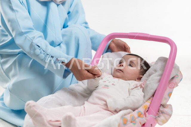 مفهوم الأمومة وتحمل المسؤولية والاهتمام بالأبناء, جلوس الرضيعة على كرسيها الخاص, بورتريه لسيدة عربية خليجيه عمانية تجلس بجانب طفلتها وترتدي الحجاب والعباءة الزرقاء, والدة سعودية تقوم بقضاء اوقات ممتعة مع الاطفال, خلفية بيضاء