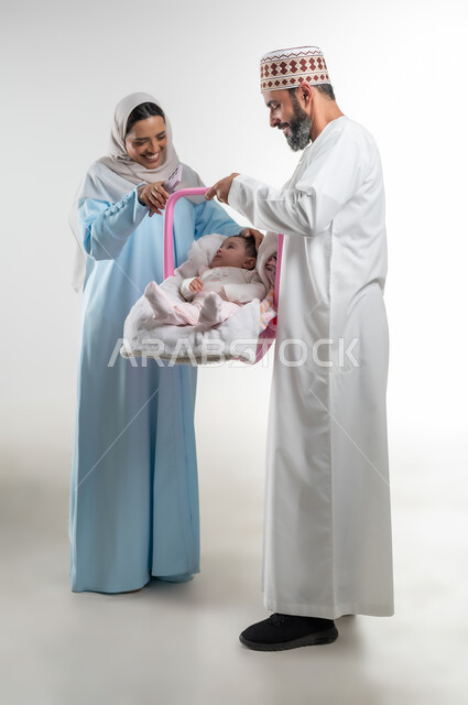 مفهوم الرعايه بالطفل, الاعتناء بالابنه ومساعدتها على الهدوء، تحمل المسؤولية والعناية بالأطفال، دور الأهل في حياه الأبناء، بورتريه من الجانب لأب عربي خليجي عماني يرتدي الدشداشة والكمة يقوم بحمل طفلته الرضيعة في كرسيها الخاص ويقف مع زوجته، خلفية بيضاء