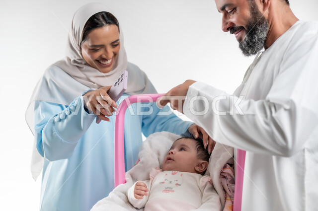 الاعتناء بالابنه ومساعدتها على الهدوء، تحمل المسؤولية والعناية بالأطفال، دور الأهل في حياه الأبناء، بورتريه من الجانب لأب عربي خليجي عماني يرتدي الدشداشة والكمة يقوم بحمل طفلته الرضيعة في كرسيها الخاص ويقف مع زوجته، مفهوم الرعايه بالطفل, خلفية بيضاء