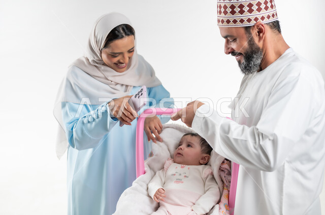 تحمل المسؤولية والعناية بالأطفال، دور الأهل في حياه الأبناء، الاعتناء بالابنه ومساعدتها على الهدوء، بورتريه من الجانب لأب عربي خليجي عماني يرتدي الدشداشة والكمة يقوم بحمل طفلته الرضيعة في كرسيها الخاص ويقف مع زوجته، مفهوم الرعايه بالطفل, خلفية بيضاء