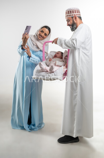 دور الأهل في حياه الأبناء، الاعتناء بالابنه ومساعدتها على الهدوء، التقاط صورة سيلفي تذكارية عبر الهاتف المحمول، بورتريه من الجانب لأب عربي خليجي عماني يرتدي الدشداشة والكمة يقوم بحمل طفلته الرضيعة في كرسيها الخاص ويقف مع زوجته، مفهوم الرعايه بالطفل, خلفية بيضاء
