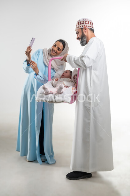 الاعتناء بالابنه ومساعدتها على الهدوء، التقاط صورة سيلفي تذكارية عبر الهاتف المحمول، دور الأهل في حياه الأبناء، بورتريه من الجانب لأب عربي خليجي عماني يرتدي الدشداشة والكمة يقوم بحمل طفلته الرضيعة في كرسيها الخاص ويقف مع زوجته، مفهوم الرعايه بالطفل, خلفية بيضاء
