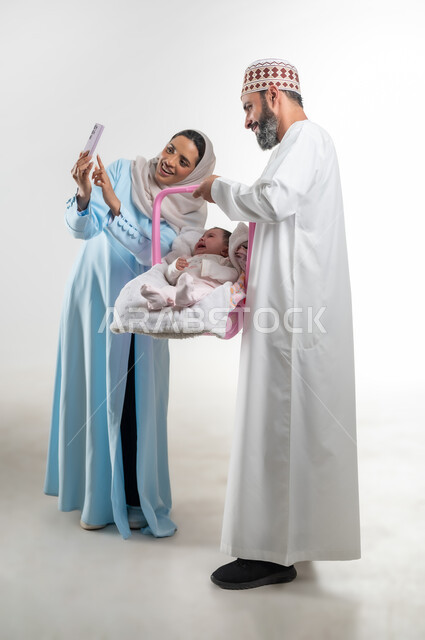 التقاط صورة سيلفي تذكارية عبر الهاتف المحمول، الاعتناء بالابنه ومساعدتها على الهدوء، دور الأهل في حياه الأبناء، بورتريه من الجانب لأب عربي خليجي عماني يرتدي الدشداشة والكمة يقوم بحمل طفلته الرضيعة في كرسيها الخاص ويقف مع زوجته، مفهوم الرعايه بالطفل, خلفية بيضاء