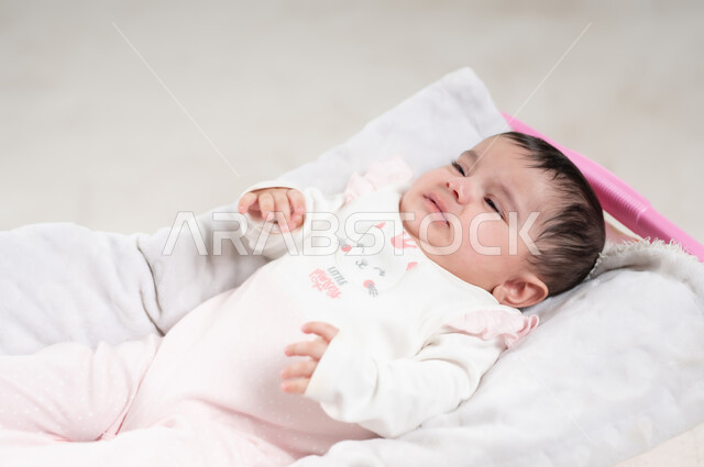 الملامح الناعمة والبريئة، مفهوم النعومة والطفولة، ايماءات البكاء والحاجة إلى شيء ما, بورتريه لطفلة عربيه خليجيه عمانية لطيفة, رضيعة سعودية ترتدي لباس قطني مستلقية على كرسي باللون الوردي، خلفية بيضاء