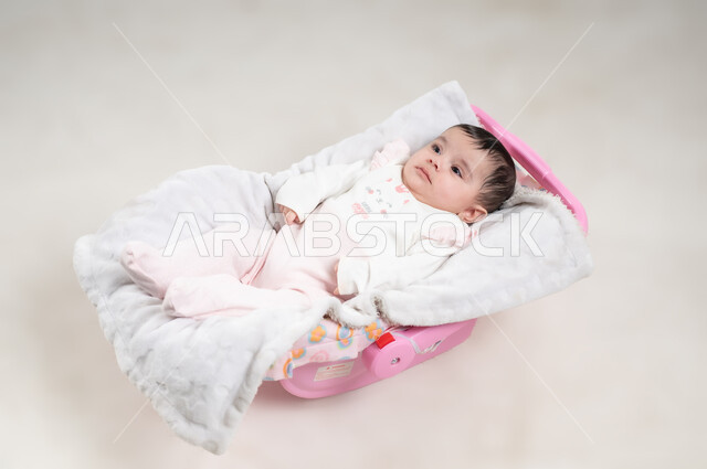 الملامح الناعمة والبريئة، رضيعة سعودية تقوم بالنظر إلى شيء ما, مفهوم البراءة و الطفولة, بورتريه  لطفلة عربيه خليجيه عمانية لطيفة ترتدي لباس قطني مستلقية على كرسي باللون الوردي, خلفية بيضاء