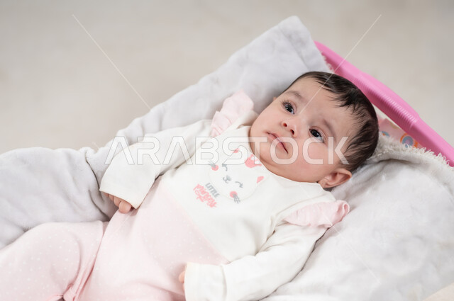 الملامح الناعمة والبريئة، رضيعة سعودية تقوم بالنظر إلى شيء ما, مفهوم البراءة و الطفولة, بورتريه  لطفلة عربيه خليجيه عمانية لطيفة ترتدي لباس قطني مستلقية على كرسي باللون الوردي, خلفية بيضاء