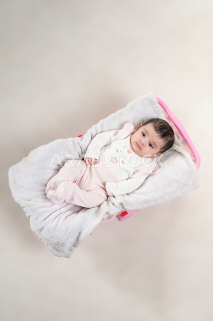 Soft and innocent features, concept of innocence and childhood, gestures of playing and looking at something, vertical overhead portrait of a cute Omani Gulf Arab girl wearing cotton clothes lying on a pink chair, white background
